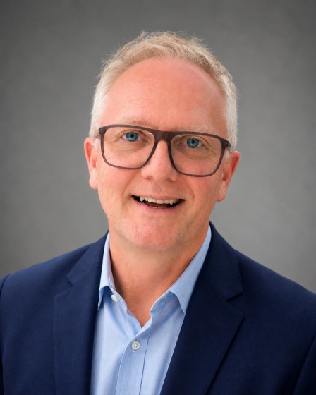 Professional headshot of a man in a navy blazer and dark-framed glasses smiling at the camera