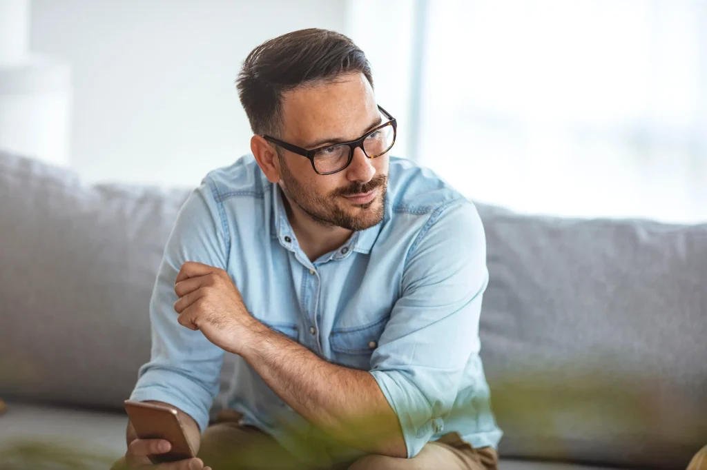 Man with beard and glasses sitting on a couch holding a smartphone, looking thoughtfully off to the side