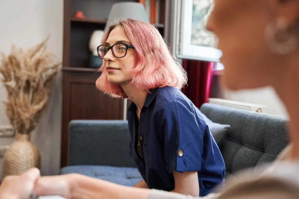 Woman with pink hair and glasses sitting attentively during a conversation