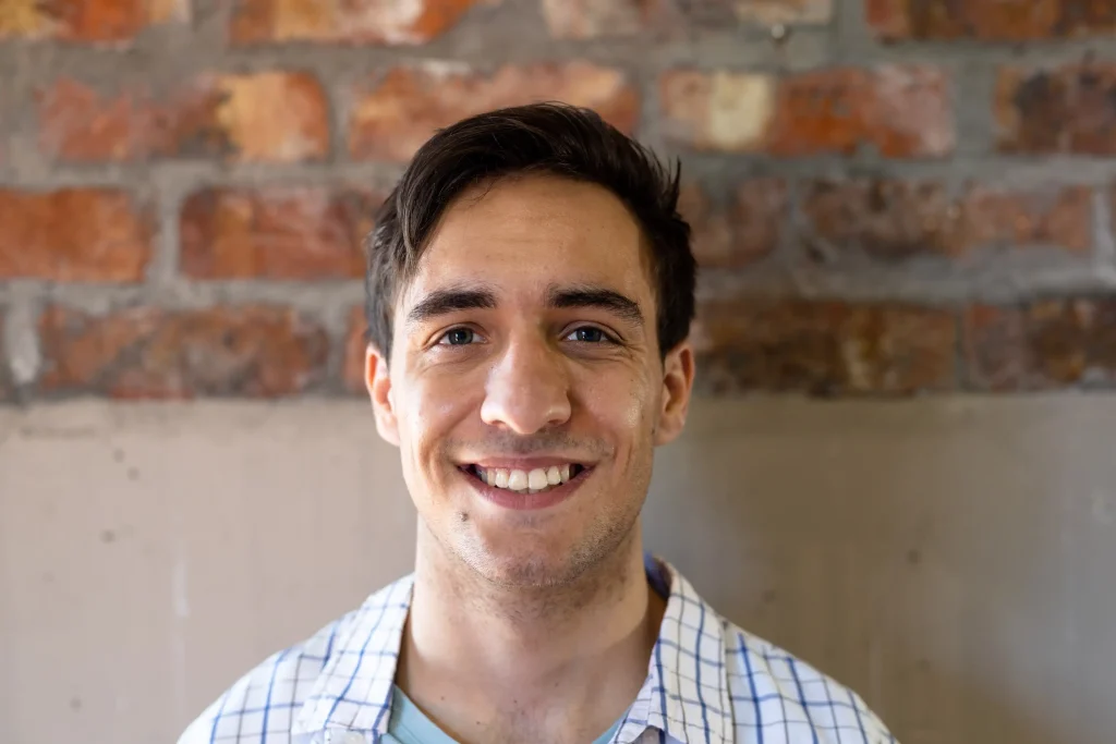 Young man in a checkered shirt smiling at the camera in front of a brick wall