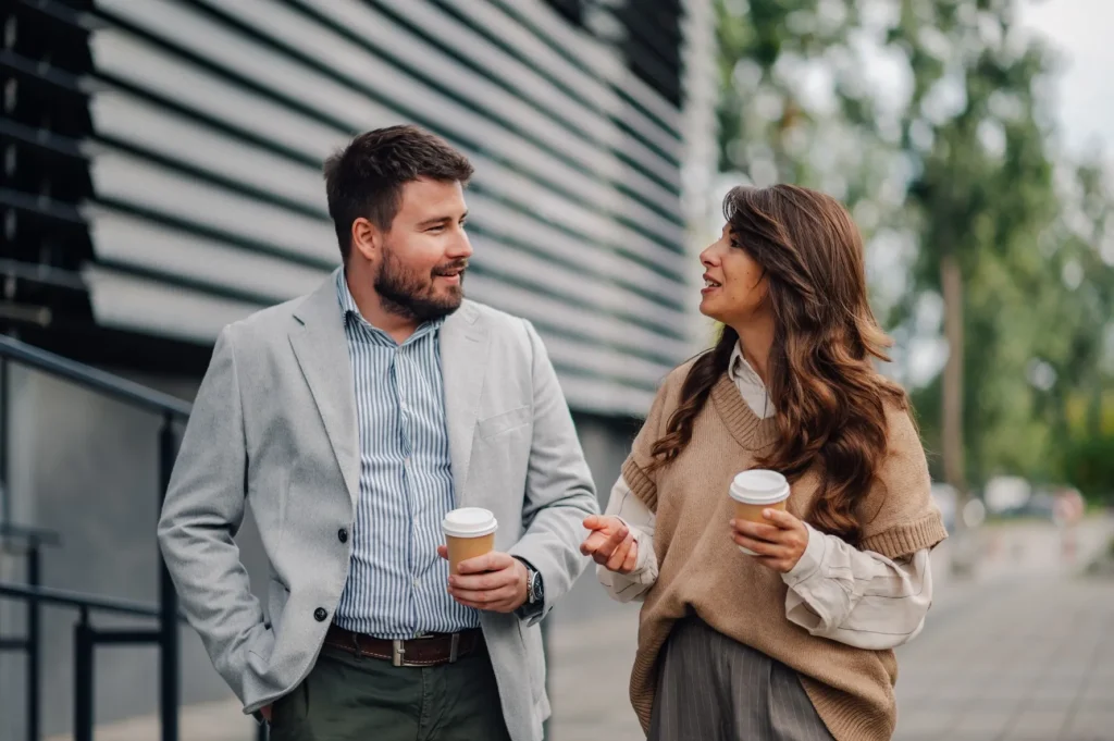 Man in a grey blazer and woman in a knit vest walking together outdoors holding takeaway coffee cups