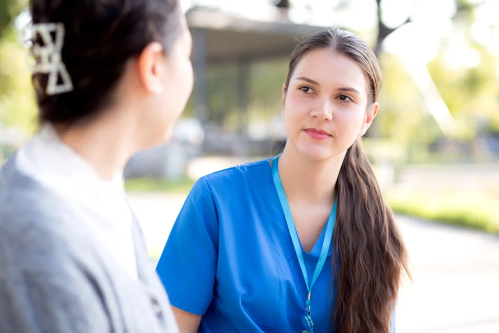 Young woman in blue medical scrubs with a lanyard talking with another woman outdoors