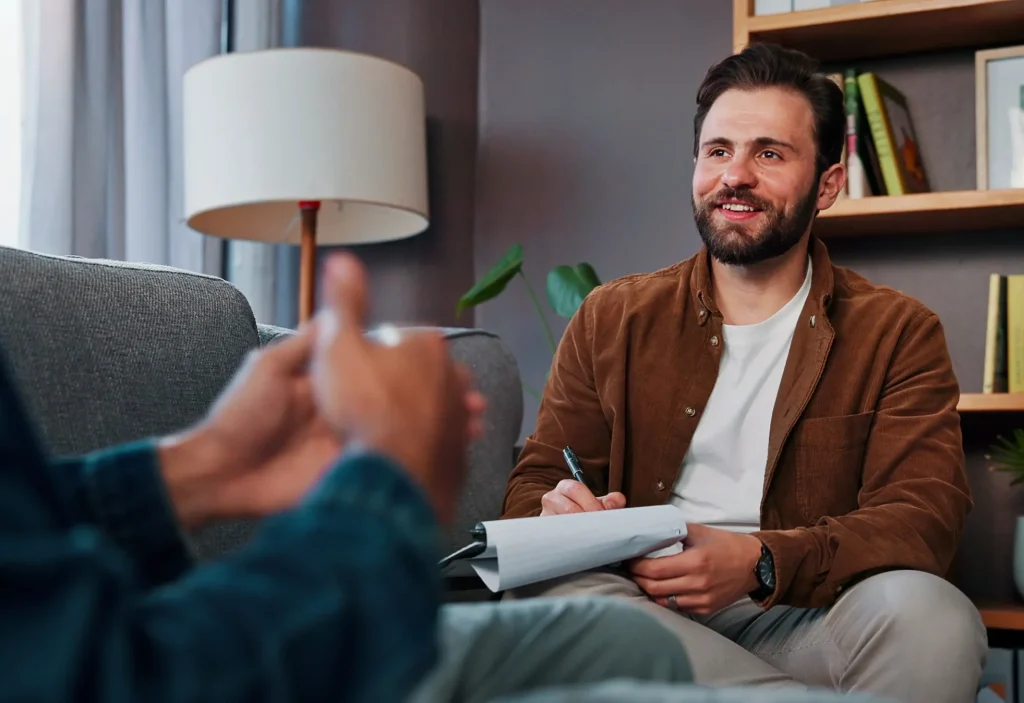 Bearded man in a brown jacket sitting on a couch holding a notepad and pen, smiling during a conversation