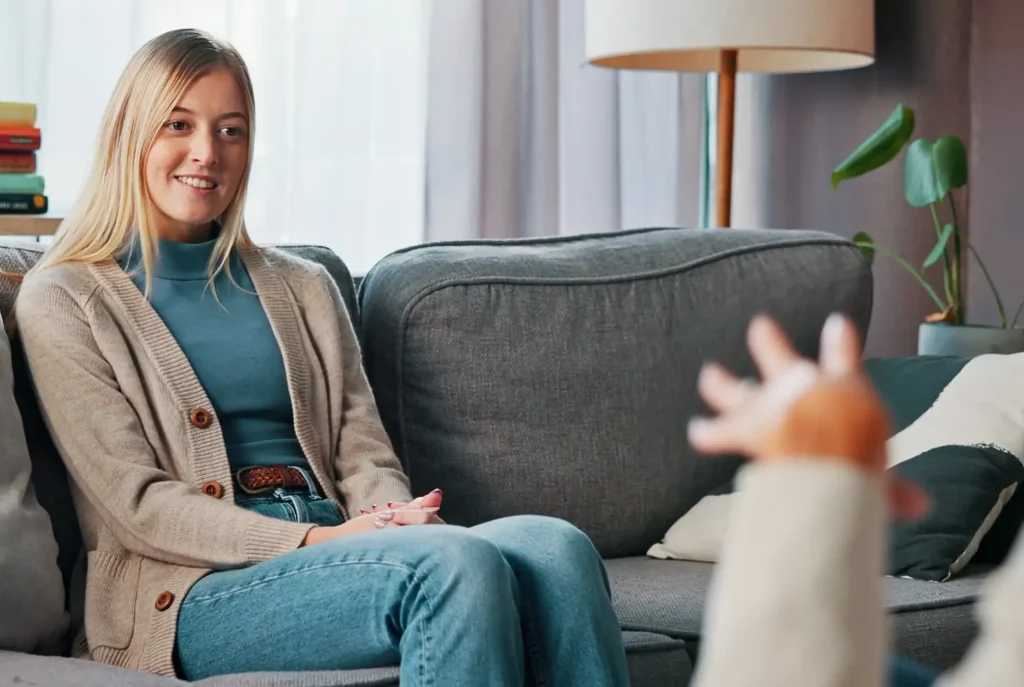 Blonde woman in a beige cardigan sitting on a grey couch smiling during a conversation