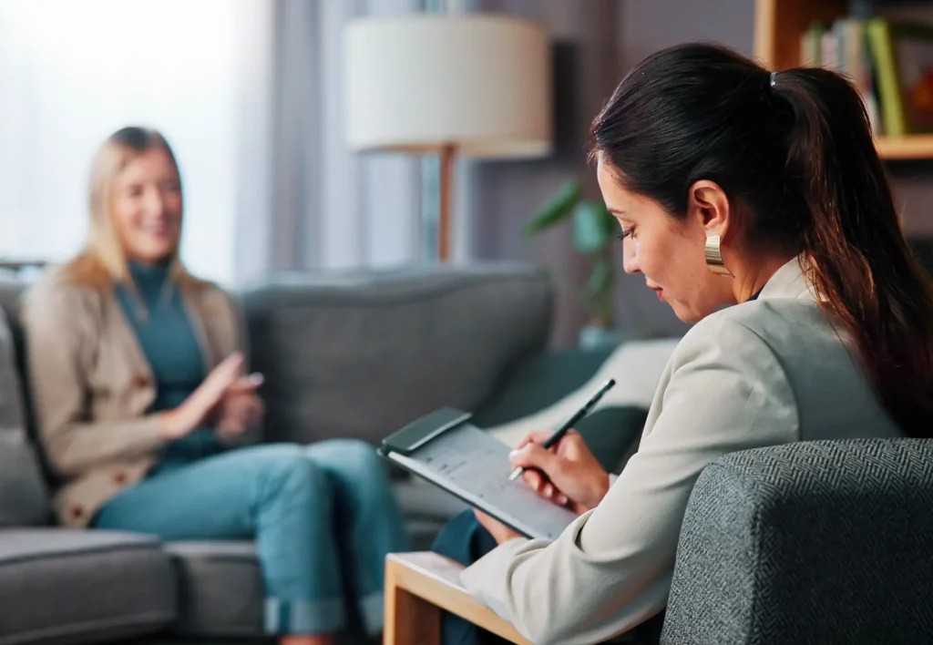 Two women in a therapy session, one sitting on a couch gesturing while the other takes notes on a tablet