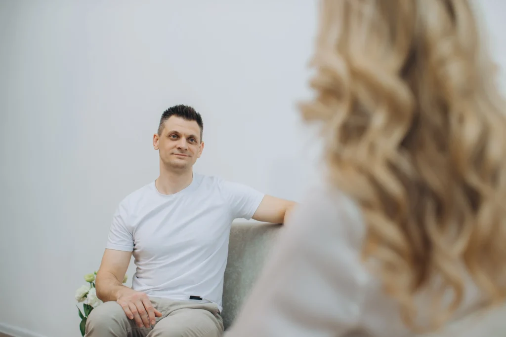 Man in a white shirt sitting on a couch looking toward a person in conversation